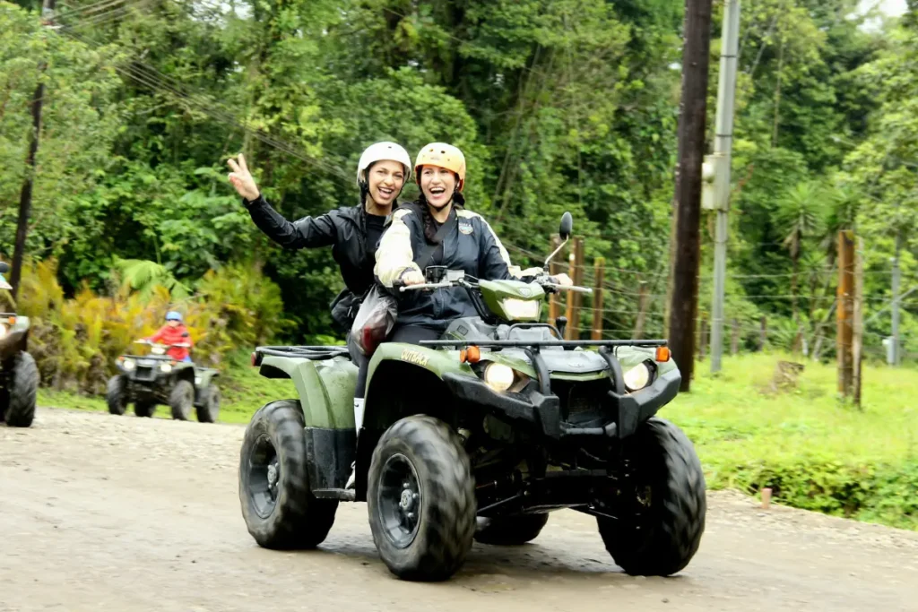 Two riders waving while driving an ATV along a rural road surrounded by rainforest in La Fortuna, Costa Rica.